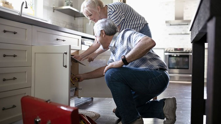 Hombre arrodillado frente al fregadero de la cocina leyendo instrucciones que sostiene una mujer. Una caja de herramientas está abierta al lado del hombre.