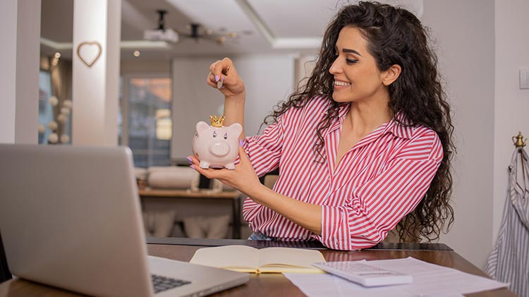 Una mujer sentada frente a unas facturas, una computadora portátil y una calculadora pone dinero en una alcancía.