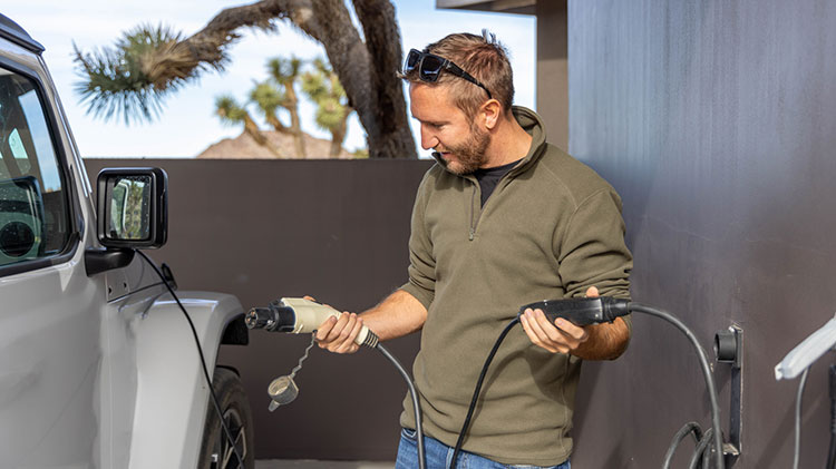 Un hombre sostiene un cargador de vehículo eléctrico junto a un vehículo blanco. 