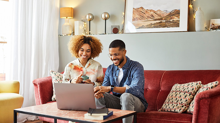 Una joven pareja sentada en un sofá color burdeos revisa su póliza de seguro de auto en su computadora portátil.