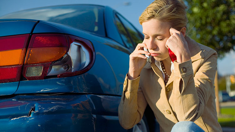 Mujer frustrada hablando por teléfono inspeccionando el daño en el parachoques del carro después de un accidente en el que no tuvo la culpa.