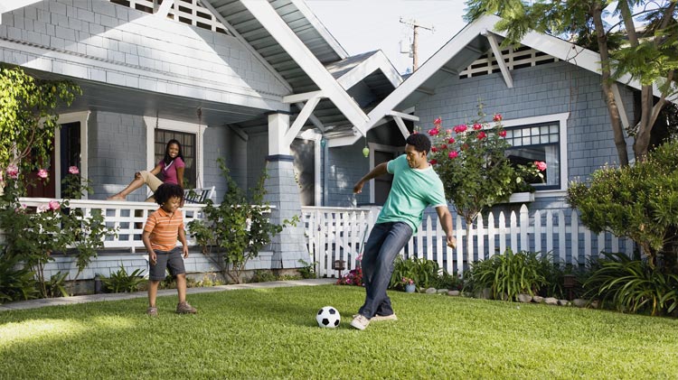 Padre e hijo jugando fútbol en el jardín delantero mientras mamá mira desde el pórtico. 